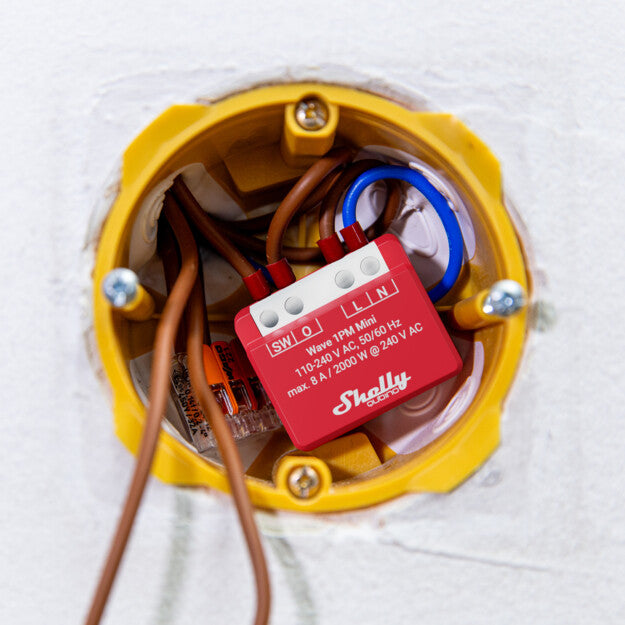 A close-up of a yellow electrical wall box featuring a red Shelly USA Shelly Wave 1PM Mini smart relay module connected to several wires inside a wall.
