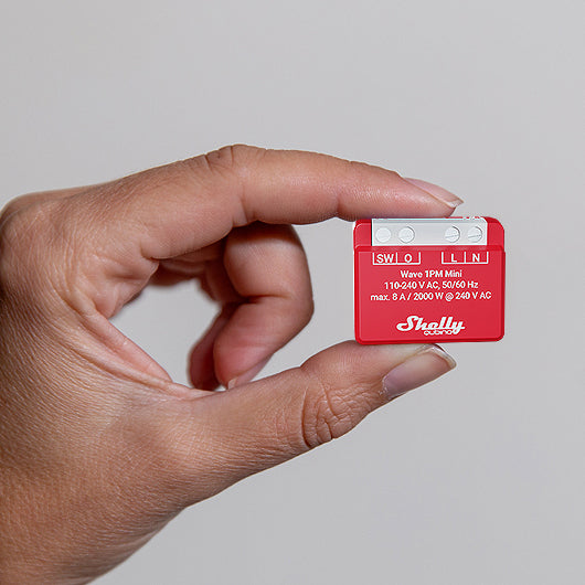 A hand holds a compact red Shelly USA Shelly Wave 1PM Mini smart relay, featuring visible white text and buttons on its surface.