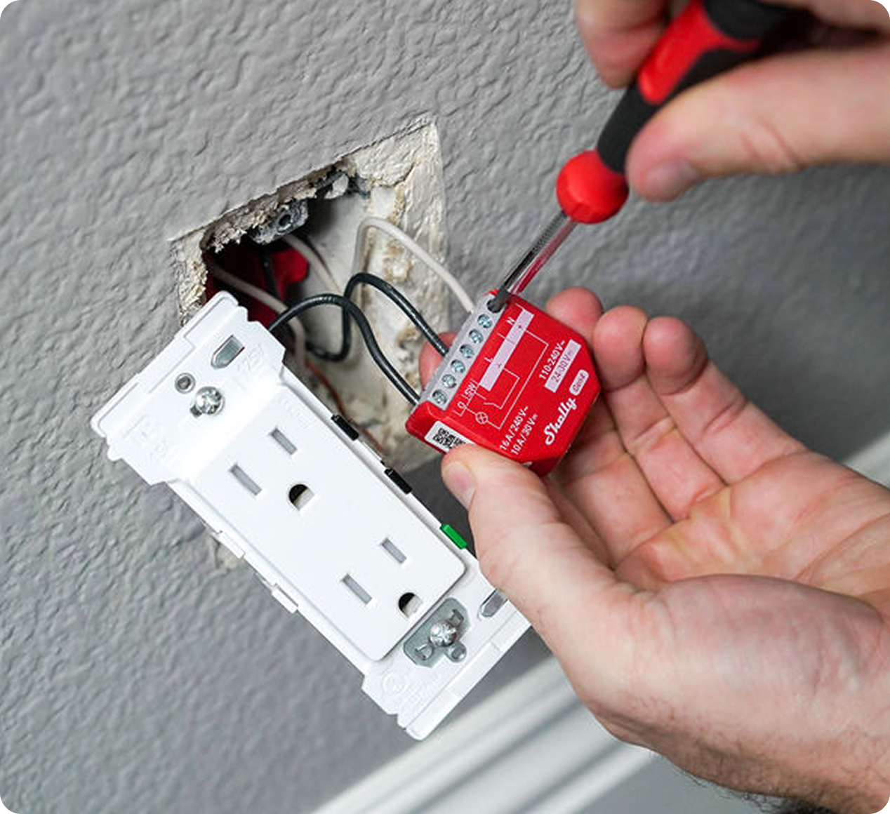 A person uses a screwdriver to install a red smart home device behind a wall power outlet, which is partially removed from the wall, revealing several connected wires.