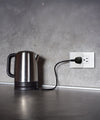 A stainless steel electric kettle sits on a gray countertop, plugged into a wall outlet with a black cord against a textured gray wall.