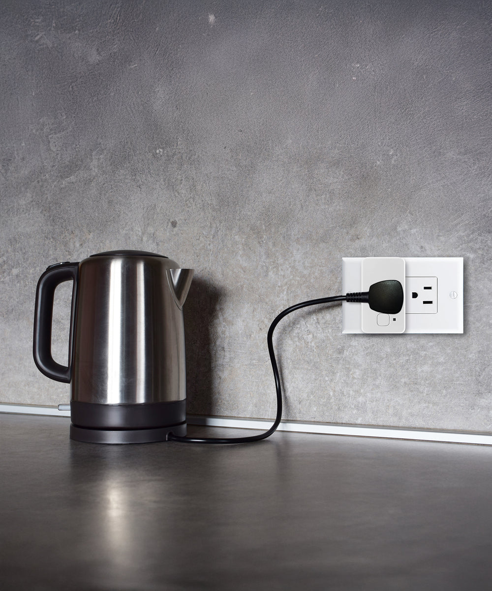 A stainless steel electric kettle sits on a gray countertop, plugged into a wall outlet with a black cord against a textured gray wall.