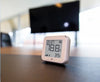 A small digital thermometer and hygrometer displays 78.8°F temperature and 39% humidity, placed on a table with a blurry TV screen and office background.