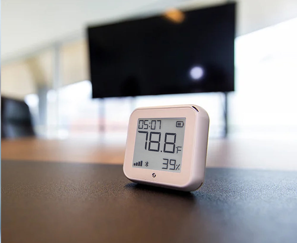 A small digital thermometer and hygrometer displays 78.8°F temperature and 39% humidity, placed on a table with a blurry TV screen and office background.