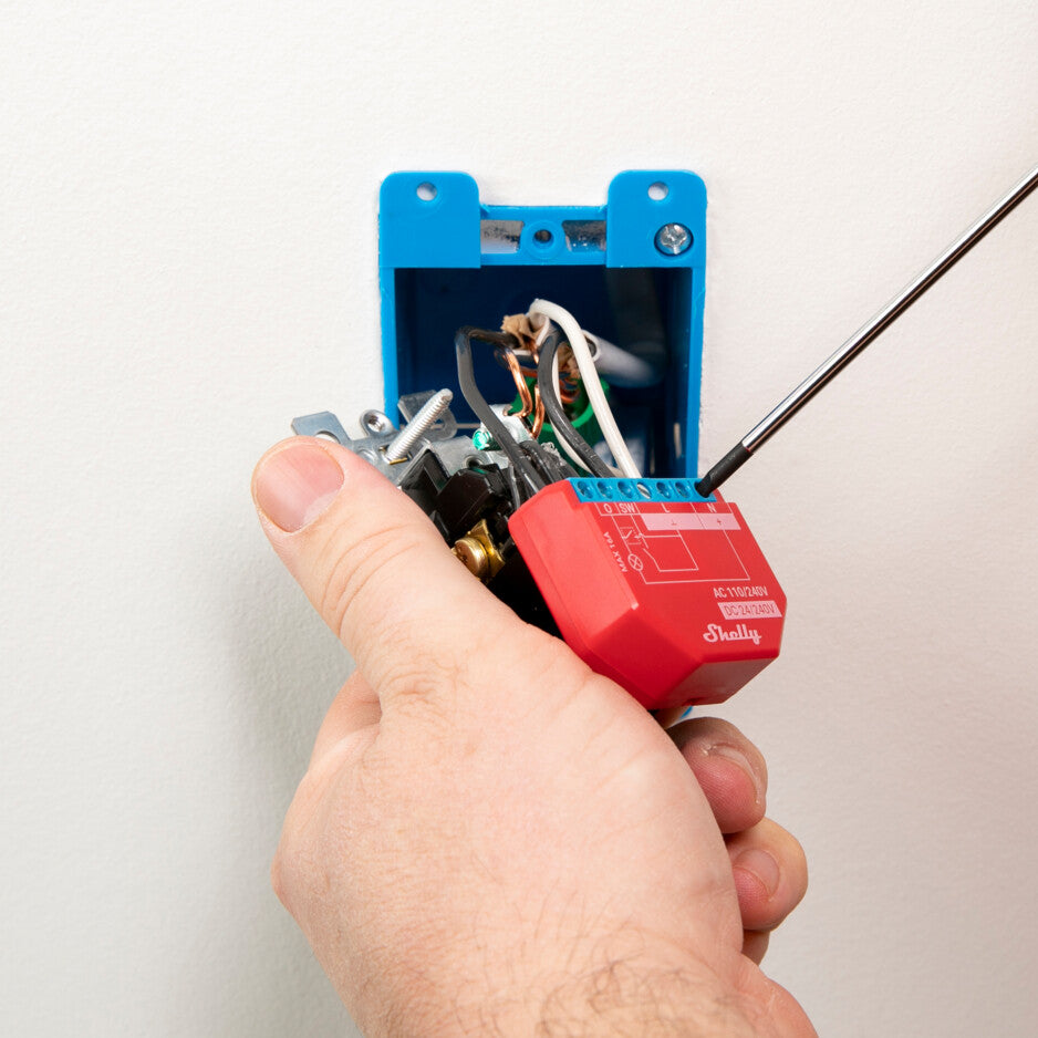 A hand is wiring a Shelly Plus 1PM Non-UL relay switch into an electrical box with several wires using a screwdriver. The box is mounted on a white wall.