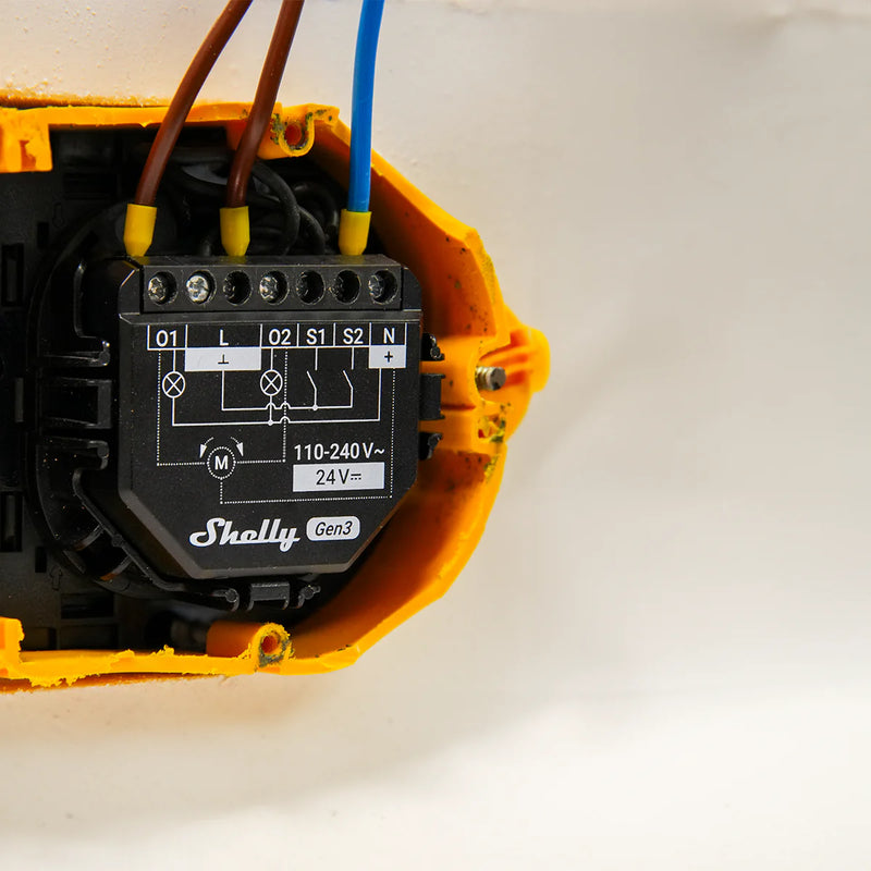 A close-up of a Shelly USA Shelly 2PM Gen3 smart switch installed in a yellow electrical box, showing labeled wiring terminals with brown and blue wires connected for accurate power metering.