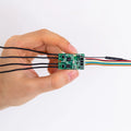 A hand holds a Shelly Uni by Shelly USA—a small green circuit board with multiple colorful wires and a smart Wi-Fi module for sensor control—set against a plain white background.
