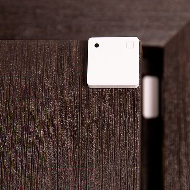 A Shelly USA Shelly Wave Door/Window LR sensor, small, white, and square, is attached near the edge of a dark wooden door with part of the hinge or door frame visible in the background.