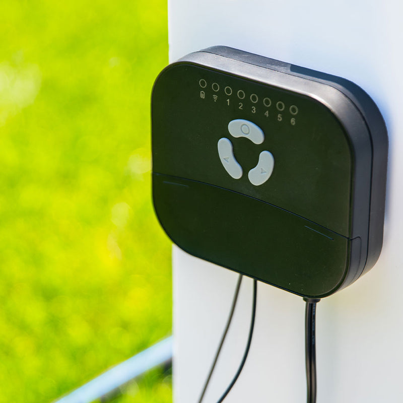 A close-up of the Shelly USA FrankEver Sprinkler Controller FK-06X, a black electronic box with buttons, indicator lights, and bottom cables, mounted on a white surface against blurred green grass.