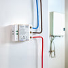 A white smart relay switch with blue, brown, and red wires connected is mounted on a white wall near an electrical panel box. The device has printed labels and indicator lights.