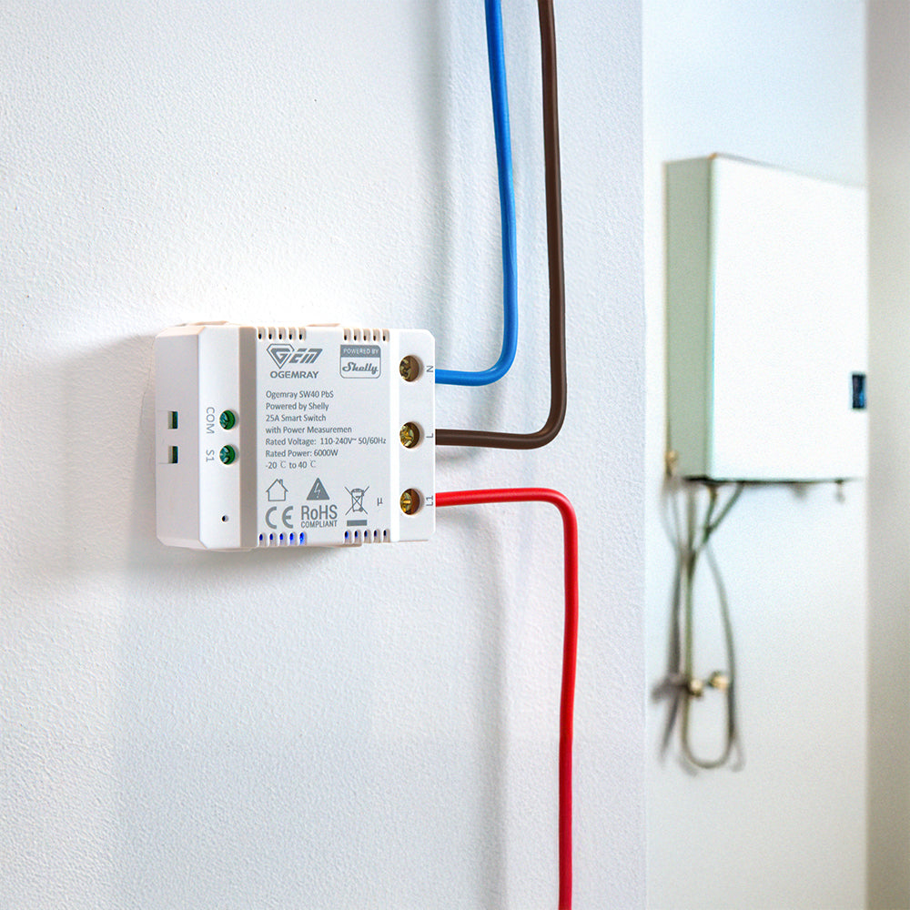 A white smart relay switch with blue, brown, and red wires connected is mounted on a white wall near an electrical panel box. The device has printed labels and indicator lights.