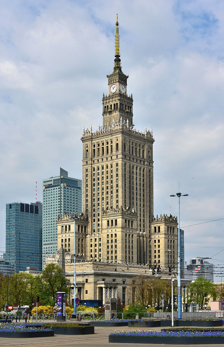 The image shows the Palace of Culture and Science, a tall historic building with a clock tower, surrounded by modern skyscrapers and trees, under a partly cloudy sky in Warsaw, Poland.