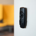 A Shelly USA Shelly BLU Remote Control ZB Black is mounted on a light gray wall, with a blurred yellow chair visible in the background.