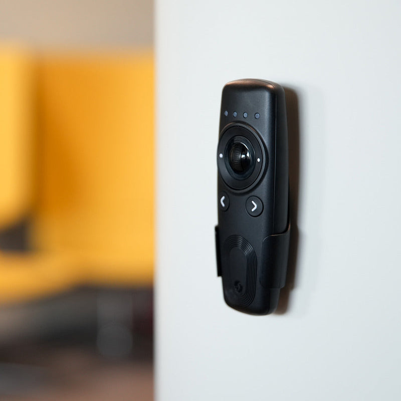 A Shelly USA Shelly BLU Remote Control ZB Black is mounted on a light gray wall, with a blurred yellow chair visible in the background.
