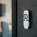 The Shelly USA Shelly BLU Remote Control ZB White with black buttons is mounted on a dark wall near a glass partition, with an office chair and desk blurred in the background.