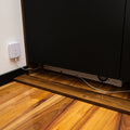 A Shelly Flood Gen4 water leak sensor is plugged into a wall outlet, with its cable leading to a black cabinet above a wooden floor. Another electronic device can be seen partially under the cabinet.