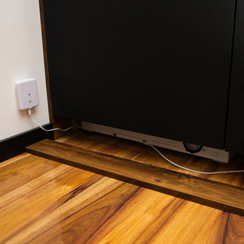 A Shelly Flood Gen4 water leak sensor is plugged into a wall outlet, with its cable leading to a black cabinet above a wooden floor. Another electronic device can be seen partially under the cabinet.