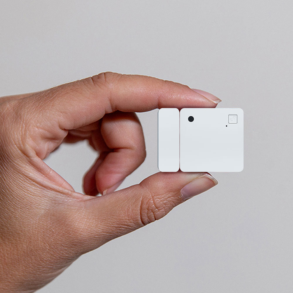 A hand holds the Shelly USA Shelly Wave Door/Window LR, a small white rectangular device with a camera lens and button, against a plain light background.