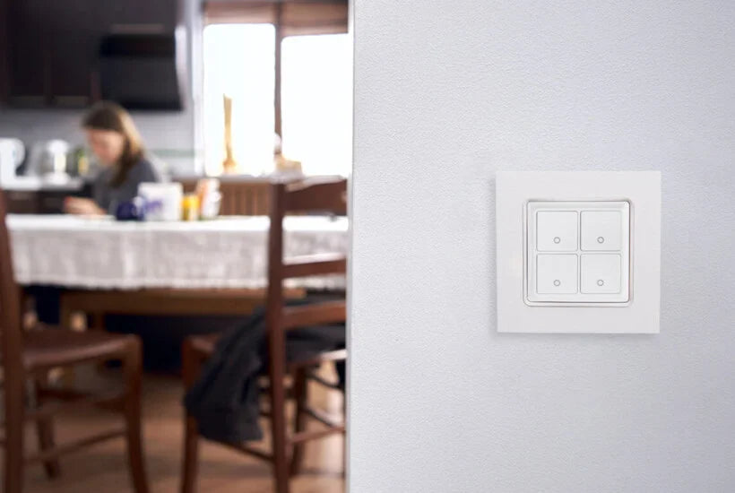 A white wall-mounted light switch is in focus on the right, while in the blurred background, a person sits at a dining table in a kitchen setting.