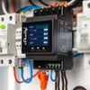A close-up of a Shelly energy meter device mounted on a panel, displaying power usage readings in watts on its screen, with various colored wires connected to terminals.