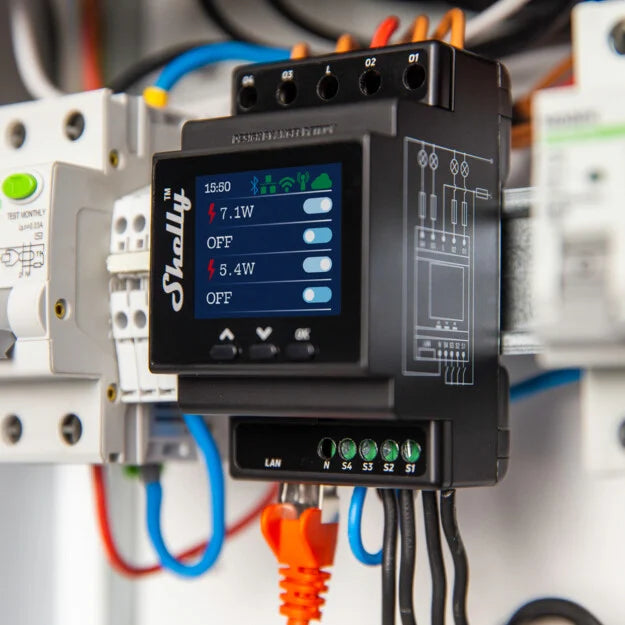 A close-up of a Shelly energy meter device mounted on a panel, displaying power usage readings in watts on its screen, with various colored wires connected to terminals.