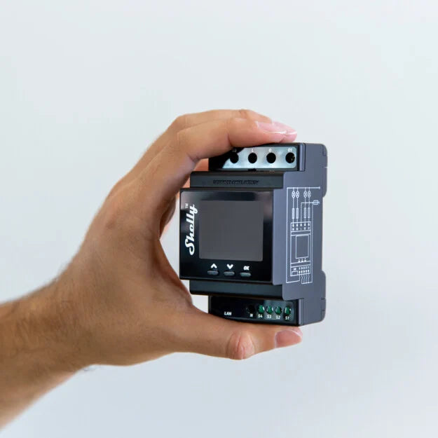 A persons hand holds a small black electronic device with a screen and labeled terminals, featuring the Shelly logo and electrical connection diagrams on one side.