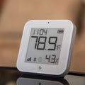 The Shelly USA Shelly BLU H&T Display ZB White shows 78.9°F and 43% humidity with digital signals and status icons on its screen, placed on a reflective surface.