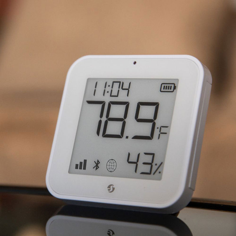 The Shelly USA Shelly BLU H&T Display ZB White shows 78.9°F and 43% humidity with digital signals and status icons on its screen, placed on a reflective surface.
