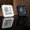 Two Shelly USA Shelly BLU H&T Display ZB White thermometers, one white and one black, show 88.1°F, 43% humidity, and the time 11:03, with their reflections visible on the glossy surface below.