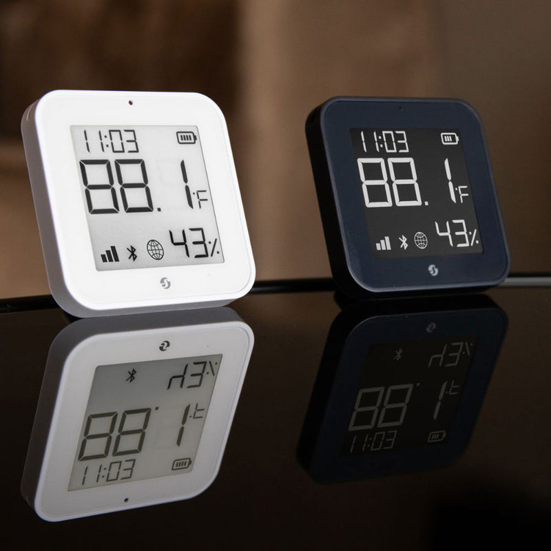 Two Shelly USA Shelly BLU H&T Display ZB White thermometers, one white and one black, show 88.1°F, 43% humidity, and the time 11:03, with their reflections visible on the glossy surface below.