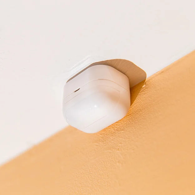 A white Apple AirPods charging case is mounted to the intersection of a white ceiling and a beige wall, resembling a security device.