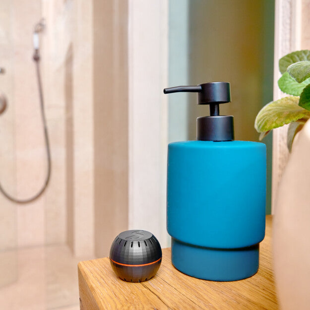 A blue soap dispenser, a small black portable speaker, and the Shelly H&T Black by Shelly USA sit on a wooden surface next to a green plant, with a glass-walled shower visible in the background.
