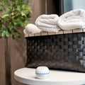 The Shelly USA Shelly H&T White, a compact round Wi-Fi humidity and temperature sensor, sits on a table in front of a black woven basket with folded white towels and a green plant in the background.