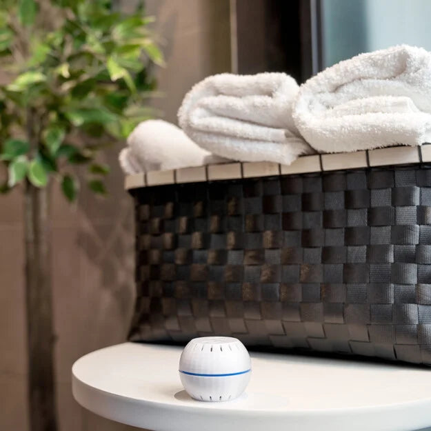 The Shelly USA Shelly H&T White, a compact round Wi-Fi humidity and temperature sensor, sits on a table in front of a black woven basket with folded white towels and a green plant in the background.