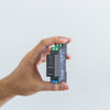 A hand holds a small black electronic device labeled Shelly with connection terminals and indicator lights on the side, against a plain light gray background.
