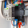 Close-up of an electrical device labeled “Shelly” with status lights for power, Wi-Fi, LAN, and two outputs, mounted on a DIN rail with various colorful connected wires.