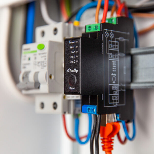 Close-up of an electrical device labeled “Shelly” with status lights for power, Wi-Fi, LAN, and two outputs, mounted on a DIN rail with various colorful connected wires.