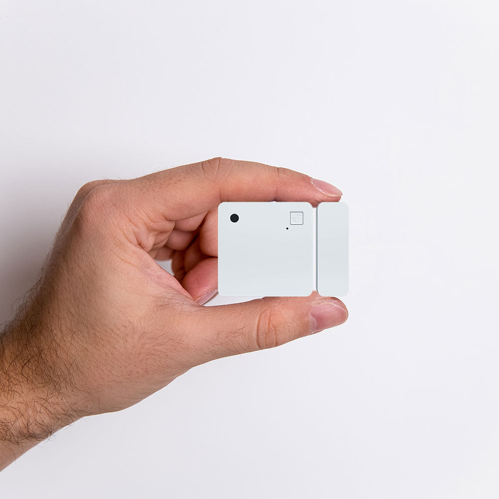 A hand holds a small, square white device with a minimalist design against a plain white background. The device has a small circular button, a tiny dot, and a rectangular notch on its surface.