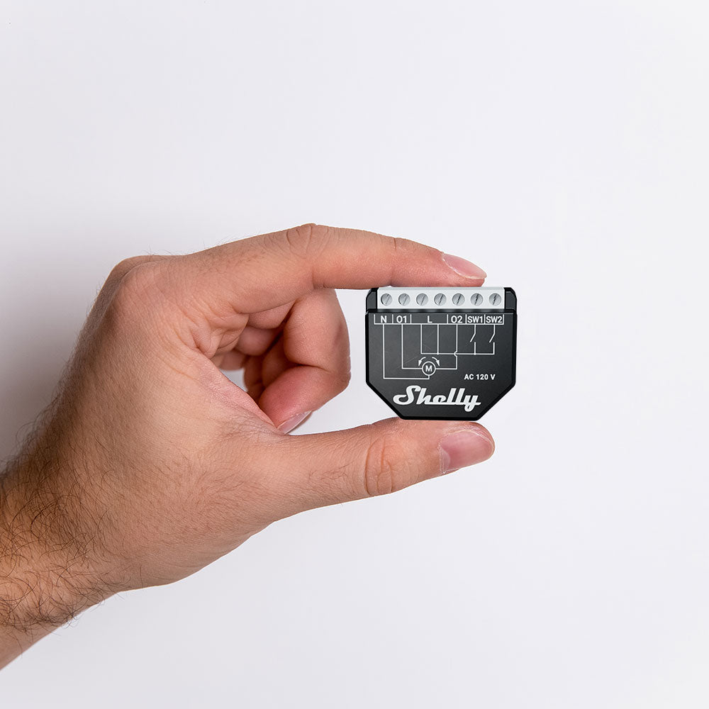 A hand holds a small black Shelly USA Shelly Wave Shutter LR device, featuring visible wiring diagrams and connection points on its top edge, set against a plain white background.