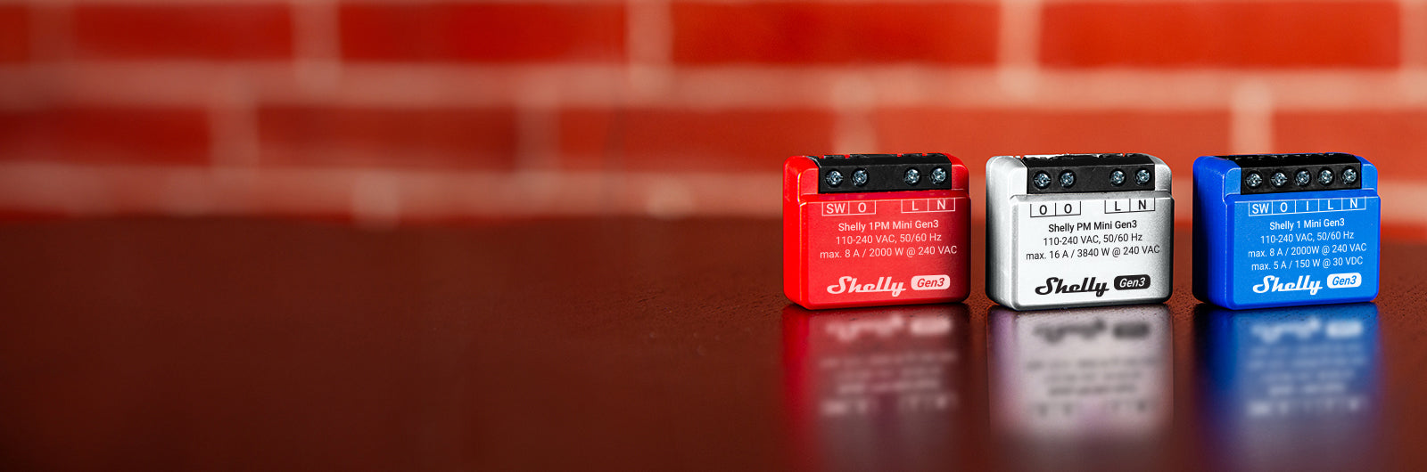 Three small Shelly Plus wifi relays in red, white, and blue sit side by side on a polished wooden surface, with a blurred red brick wall in the background.