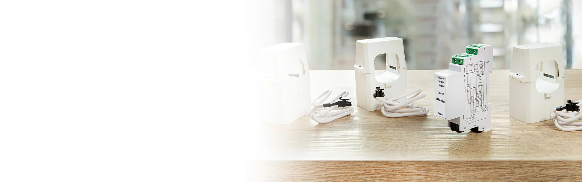A white electronic device with a circuit diagram and green terminal blocks stands on a wooden surface, surrounded by three white current transformers with coiled wires.