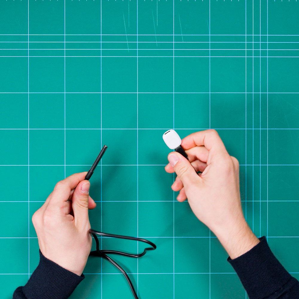A person holds a Shelly Temp. Sensor DS18B20 3.5mm jack by Shelly Europe in one hand and a white USB connector in the other over a green cutting mat with a grid pattern.