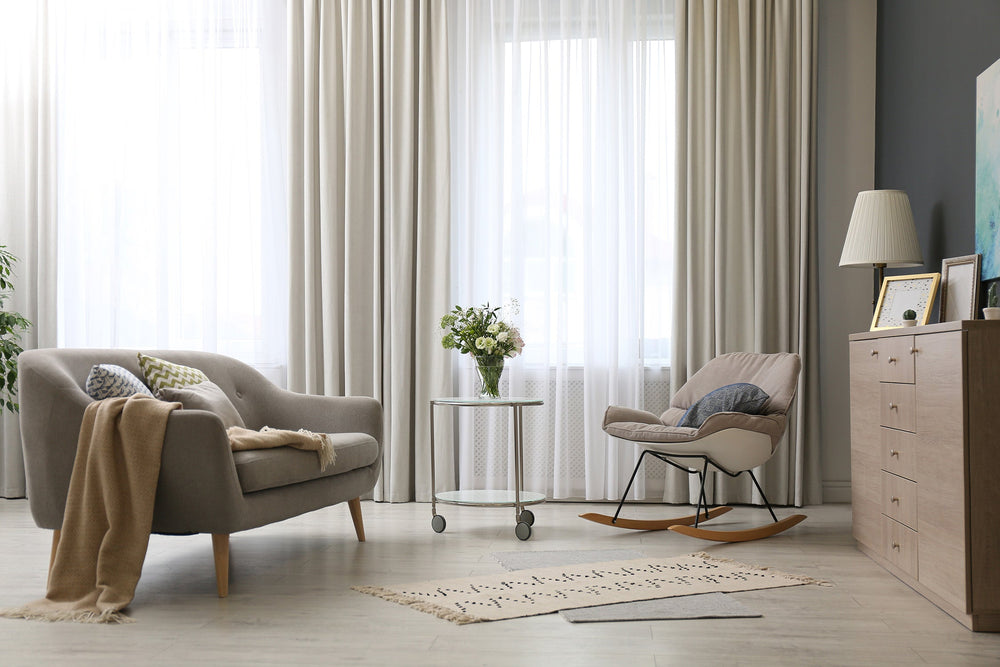 A cozy living room with a gray sofa, beige blanket, round glass coffee table with flowers, rocking chair, dresser with lamp and picture frames, and large windows with sheer white curtains.