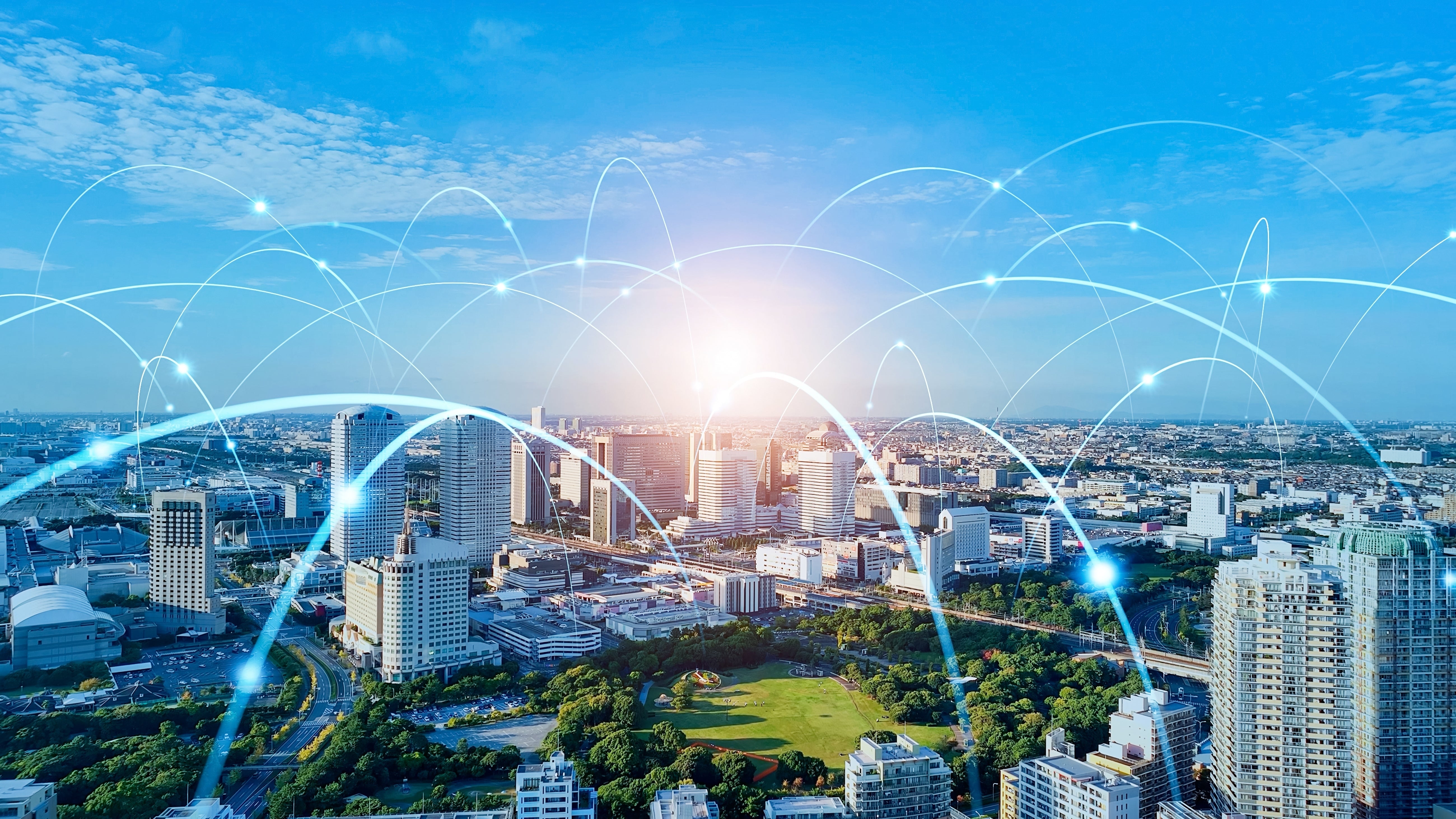 A cityscape with tall buildings and a park, overlaid with glowing arcs and digital light points, symbolizing a connected smart city network under a clear blue sky.