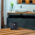 The Shelly Shelly BLU H&T ZB Black, a square device with textured lines and a central button, rests on a wooden surface in a modern bathroom featuring a sink, counter, mirror, and vase of flowers in the background.