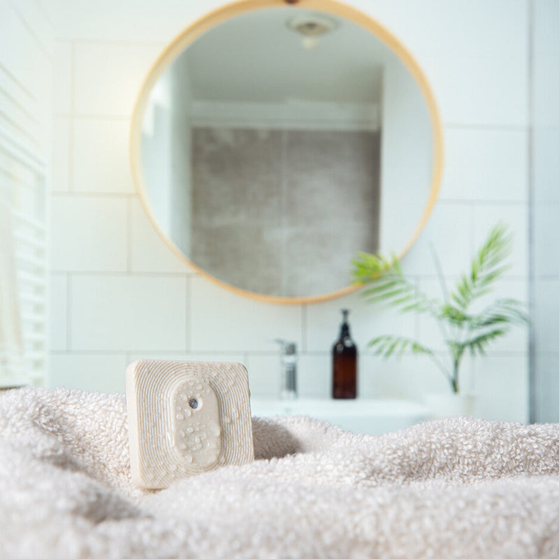 The Shelly Europe Shelly BLU H&T ZB Ivory is displayed on a soft beige towel in a bright bathroom, with a round mirror, sink, potted plant, and brown bottle softly blurred in the background.