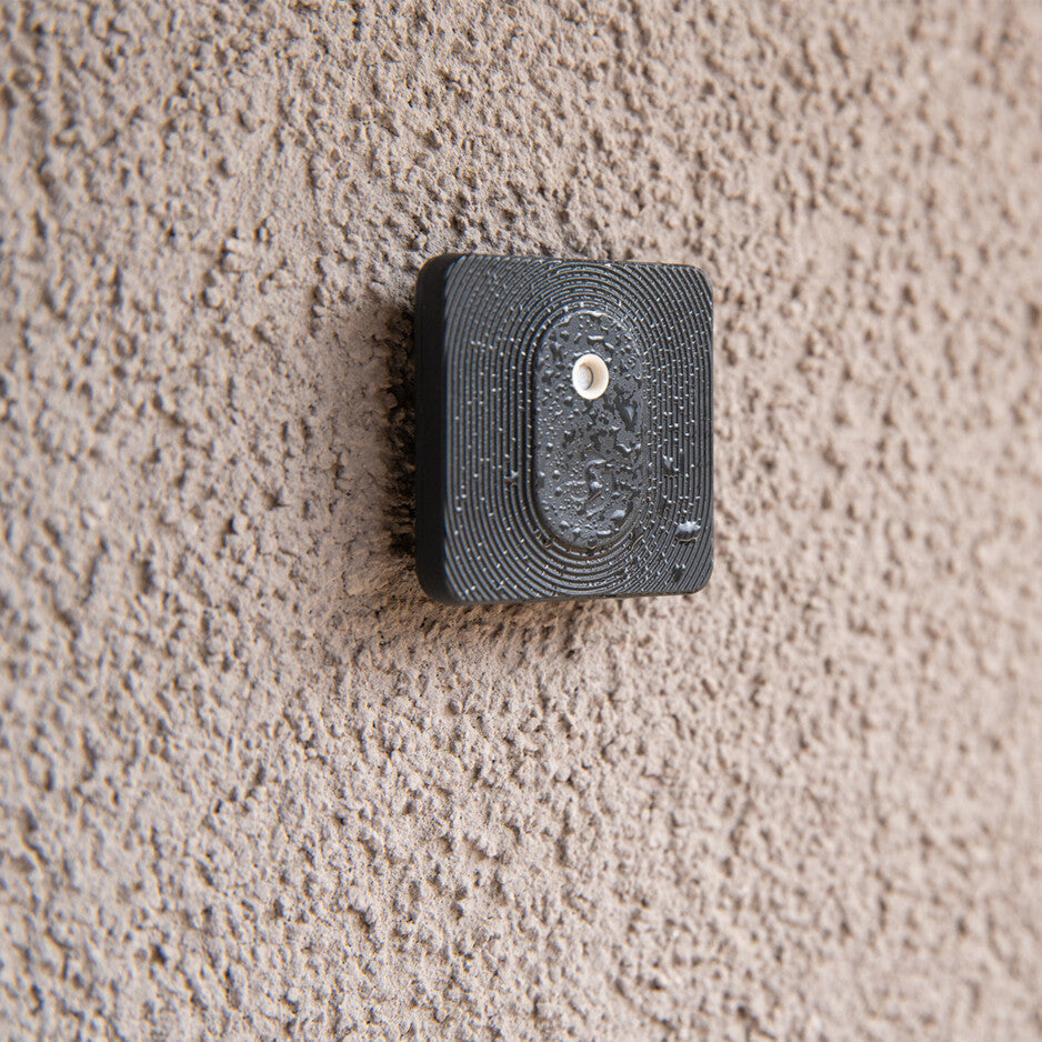 A Shelly Shelly BLU H&T ZB Black device with a white center is mounted on a textured beige wall, its surface dotted with water droplets.