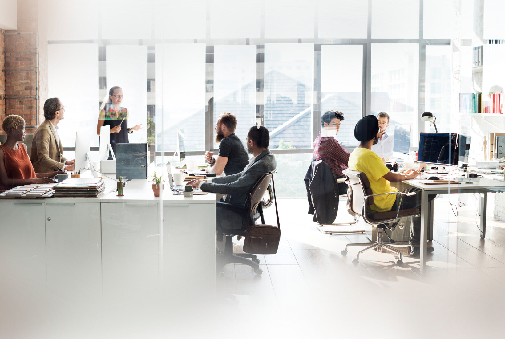 A group of people work and collaborate in a bright modern office with large windows, desks, and computers. One person stands and presents while others listen or work at their computers.