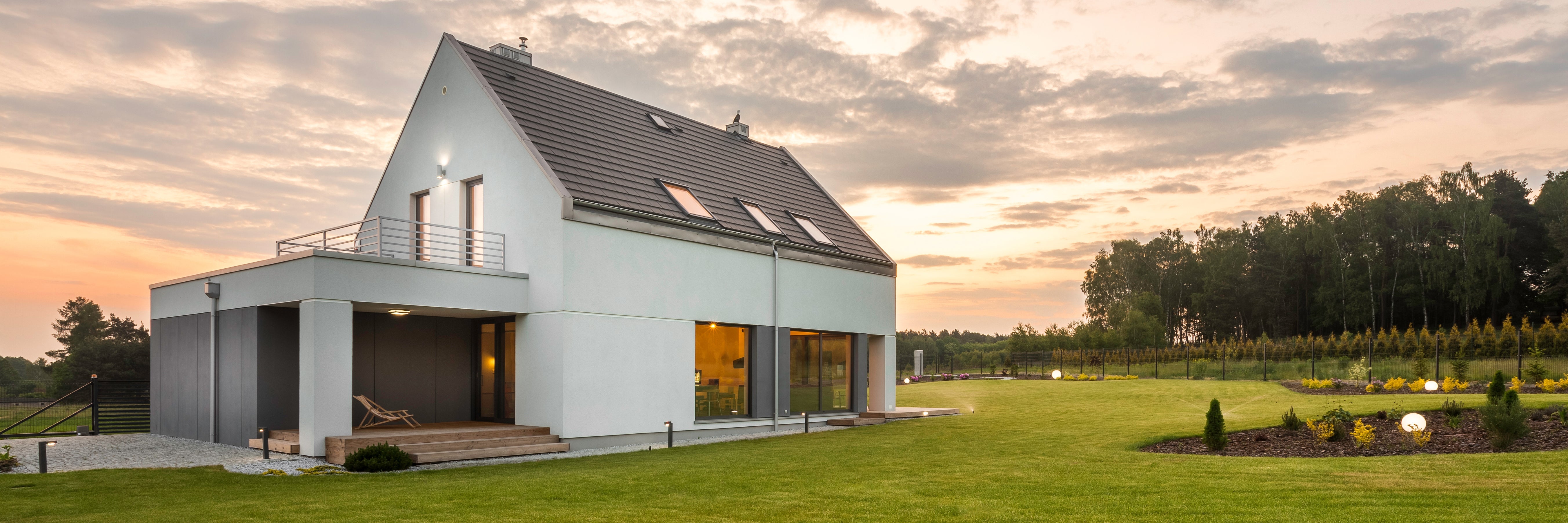 Modern two-story house with large windows, a balcony, and sloped roof, surrounded by a spacious green lawn and landscaped garden, at sunset with a tree line in the background.