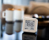 A digital thermometer and hygrometer displaying 78.8°F and 39% humidity sits on a reflective table. In the blurred background, there are two mugs and a sofa.