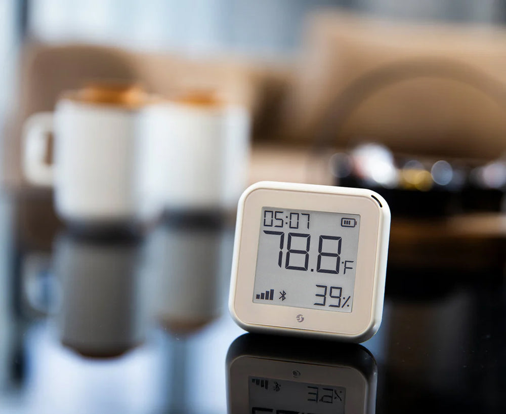 A digital thermometer and hygrometer displaying 78.8°F and 39% humidity sits on a reflective table. In the blurred background, there are two mugs and a sofa.